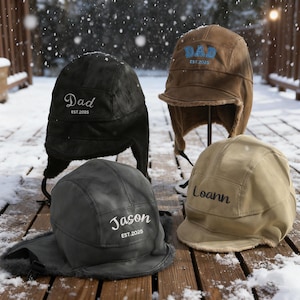 May include: Four personalised winter hats with ear flaps are displayed on a snowy wooden deck. The hats are black, brown, grey, and tan. Each hat has a name embroidered on it, with the text "Dad", "Jason", and "Loann" visible. The text "EST.2025" is also embroidered.