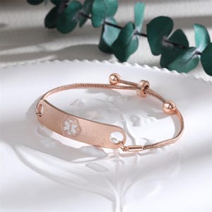 May include: Rose gold medical alert bracelet with a textured ID plate. The plate features a white medical symbol. The bracelet has a snake chain design and adjustable closure with two small spheres. The bracelet is displayed on a white surface.