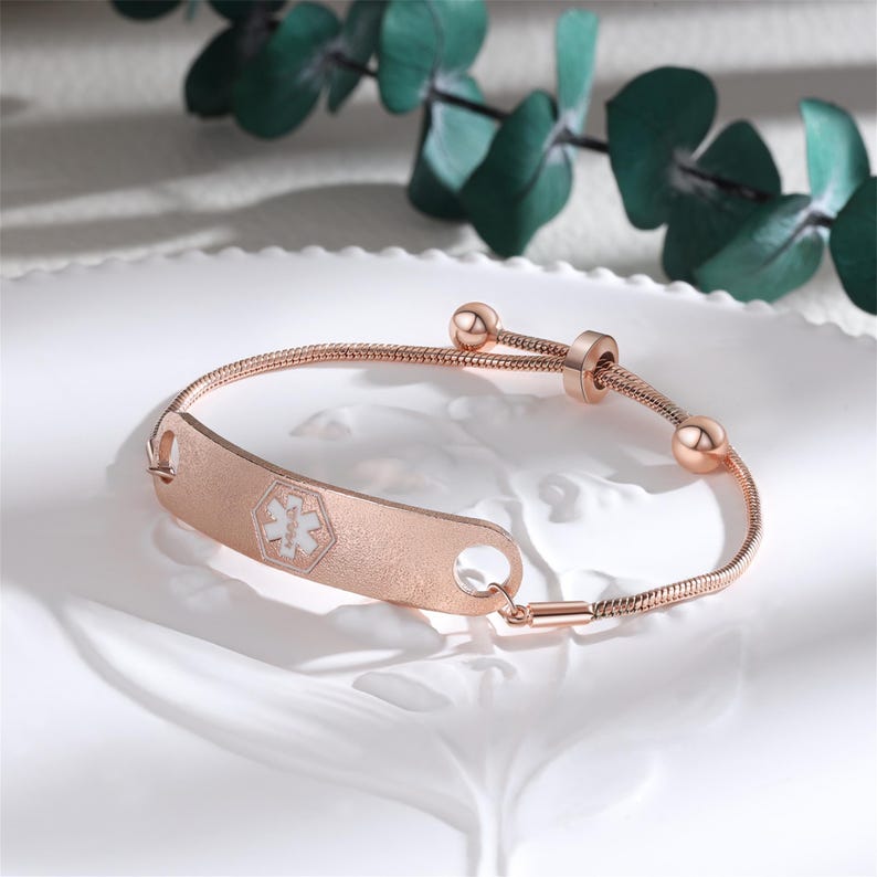 May include: Rose gold medical alert bracelet with a textured ID plate. The plate features a white medical symbol. The bracelet has a snake chain design and adjustable closure with two small spheres. The bracelet is displayed on a white surface.
