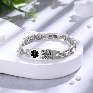 Op de afbeelding: Een zilverkleurige medische ID-armband. De armband heeft een centrale rechthoekige plaat met een zwart medisch symbool, een QR-code en de tekst "HEALTH INFO SCAN ME". De schakels van de armband hebben hartvormige accenten en heldere stenen.