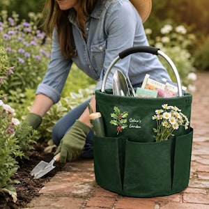 Könnte beinhalten: Eine dunkelgrüne, zylindrische Gartentasche mit einem silbernen Metallgriff und mehreren Außentaschen. Die Tasche ist mit einem aufgedruckten Stechpalmenzweig und dem Text "Selina's Garden" versehen. Sie enthält eine Handschaufel und Samentüten.
