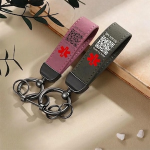 May include: Two medical alert keychains with a loop design, one in mauve and one in dark grey. Each features a red Star of Life symbol, a QR code, and the text "SCAN ME" and "HEALTH INFO" printed on the strap, attached to black metal key rings.