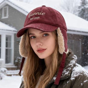 May include: A burgundy trapper hat with ear flaps and a curved brim. The hat has the word "Bonnie" embroidered on the front, along with "EST.2025". The ear flaps are lined with faux fur. Burgundy straps hang down from the ear flaps.