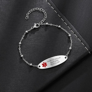 May include: Silver-toned medical ID bracelet with a chain and bead design. The oval plate features a red medical symbol and the text "DIABETES INSULIN WARFARIN PENICILLINP". The bracelet is displayed on a black surface.