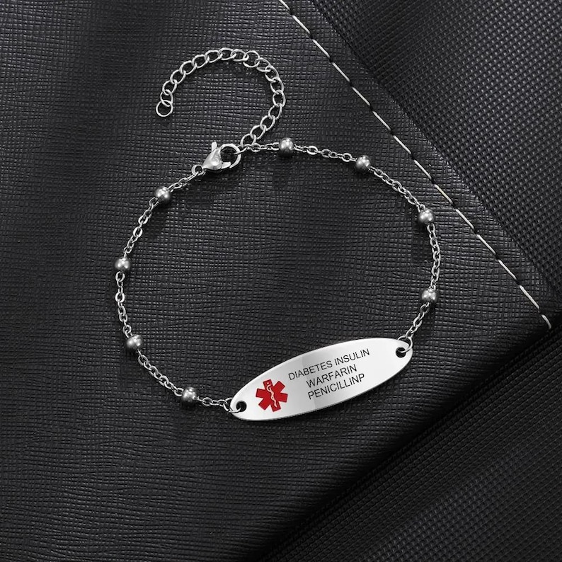 May include: Silver-toned medical ID bracelet with a chain and bead design. The oval plate features a red medical symbol and the text "DIABETES INSULIN WARFARIN PENICILLINP". The bracelet is displayed on a black surface.