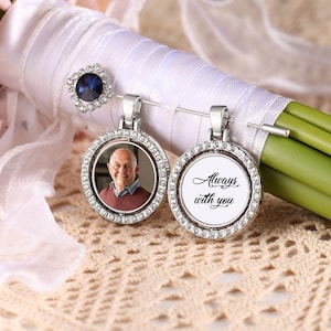 Puede incluir: Dos colgantes conmemorativos plateados, uno con una foto y otro con el texto "Always with you". Cada colgante tiene un diseño circular con un borde de cristal. También se ve un broche de cristal azul.