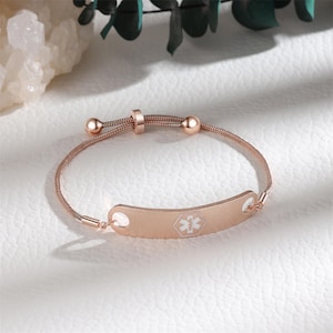 May include: Rose gold medical ID bracelet with a delicate chain and adjustable clasp. The ID plate features a white medical symbol. The bracelet is designed for safety and style, suitable for everyday wear.