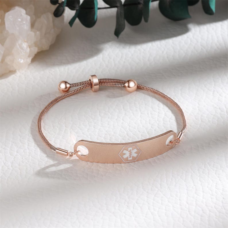 May include: Rose gold medical ID bracelet with a delicate chain and adjustable clasp. The ID plate features a white medical symbol. The bracelet is designed for safety and style, suitable for everyday wear.