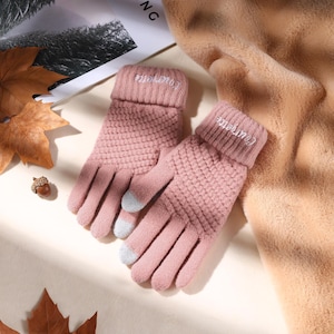 Könnte beinhalten: Ein Paar rosa Strickhandschuhe mit strukturiertem Muster. Die Bündchen sind umgeschlagen und mit dem Wort "Courgette" in Weiß bestickt. Die Fingerspitzen haben graue Pads für die Touchscreen-Bedienung. Ein beiger Schal und Herbstblätter sind im Hintergrund.