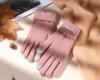 Può includere: Un paio di guanti rosa a maglia con un motivo strutturato. I polsini sono ripiegati e ricamati con la parola "Courgette" in bianco. Le punte delle dita hanno cuscinetti grigi per l'uso touchscreen. Una sciarpa beige e foglie autunnali sullo sfondo.