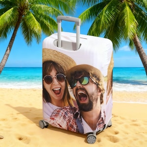 May include: White rolling suitcase featuring a custom photo print of a smiling couple in sunglasses and straw hats. The suitcase has a retractable handle and is set against a beach backdrop with palm trees and blue water.