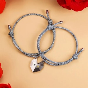 Puede incluir: Dos pulseras de amistad a juego con cordones trenzados grises y blancos. Cada pulsera tiene un dije en forma de corazón, uno blanco con una "N" negra y el otro negro con una "M" amarilla. Las pulseras están conectadas por un nudo.