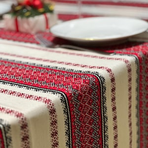 Op de afbeelding: Close-up van een crèmekleurig tafelkleed met een rood en zwart geometrisch patroon. Een wit bord, vork en wijnglas staan op het kleed, wat een feestelijke eetsfeer creëert.