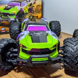 Puede incluir: Un monster truck teledirigido verde neón y morado con neumáticos negros grandes. El camión tiene un capó negro con la palabra "Beast" y un gráfico de toro. El parabrisas es rosa. El fondo muestra un camión similar.