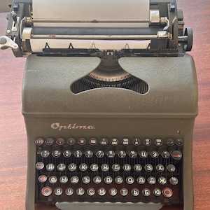 以下が含まれることがあります： 白い紙が挿入されたオリーブグリーンのOptimaタイプライター。キーボードは、白い文字と赤いアクセントが付いた黒いキーを備えています。「Optima」の文字がタイプライターの前面に印刷されています。