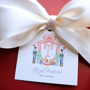 May include: A red gift box tied with a cream-colored satin bow. A white tag with a watercolor design and the words "Merry Christmas" is attached. The tag features a festive illustration with nutcrackers and a monogram.