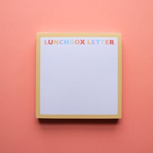 May include: A square notepad with a yellow border and the words "LUNCHBOX LETTER" in colorful letters across the top. The notepad is on a coral-colored background.
