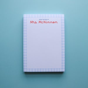 May include: A rectangular notepad with a light blue gingham border and a light blue interior. The text "FROM THE DESK OF" and "Mrs. McKinnon" are printed in red cursive. The notepad is on a light blue background.