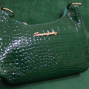 Peut inclure: Un sac à main vert émeraude brillant avec une texture peau de crocodile. Le sac a une forme incurvée, une fermeture éclair et des détails métalliques dorés. Le nom de la marque "Sunnypetty" est affiché en lettres dorées.