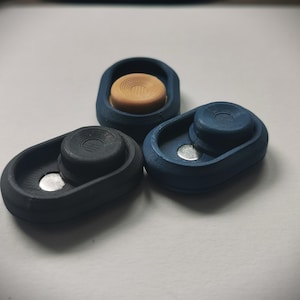 Puede incluir: Tres pequeños objetos de forma ovalada, impresos en 3D, con una hendidura circular en la parte superior. Los objetos son negros, azules y marrones.