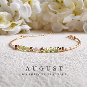 May include: A rose gold bracelet with a textured band, featuring peridot and pearl beads. The bracelet is labelled "AUGUST BIRTHSTONE BRACELET" in a stylish font, set against a soft, neutral background with white flowers.