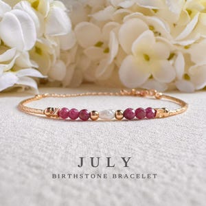 May include: A delicate gold-toned bracelet featuring a row of vibrant red gemstones, a white pearl, and gold beads. The bracelet is labeled "JULY BIRTHSTONE BRACELET" in a soft, elegant font. The background includes white flowers.