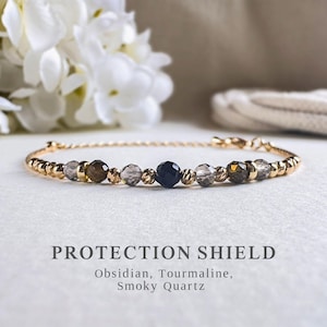 May include: A gold bracelet with a central dark blue obsidian bead, flanked by smoky quartz and tourmaline beads. The bracelet includes small gold beads. The image text reads "PROTECTION SHIELD" and lists the stones used in the bracelet.
