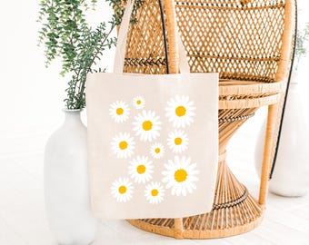 May include: A white canvas tote bag with a design of yellow and white daisies. The bag is sitting on a wicker chair.