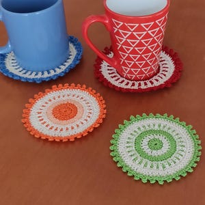 May include: Four crocheted coasters in different colors. The coasters are round and have a white center with a colorful outer ring. The coasters are on a wooden table.