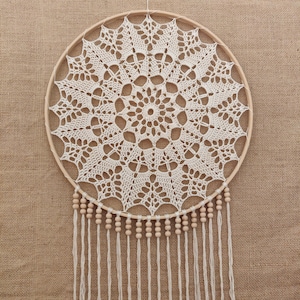 Pode incluir: Um apanhador de sonhos de crochê branco com contas de madeira penduradas na parte inferior. O apanhador de sonhos é circular e tem um design delicado e intrincado.