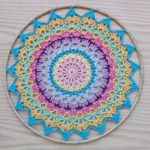 Op de afbeelding: Een handgemaakte, kleurrijke gehaakte dromenvanger. Het cirkelvormige ontwerp heeft concentrische ringen in pastelkleuren, waaronder roze, geel, blauw en paars. De buitenste ring is van natuurlijke houtkleur en heeft blauwe driehoekaccenten.