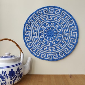 May include: A blue and white crocheted round rug with a geometric pattern. The rug has a white centre with a blue border and a blue and white geometric pattern around the edge.