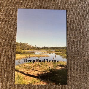 May include: A postcard featuring a scenic landscape of Deep East Texas. The image shows a body of water surrounded by green grass and trees under a clear blue sky. The text "Deep East Texas" is printed in black.