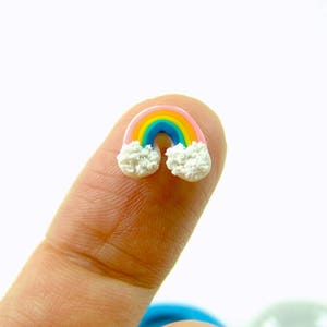 Puede incluir: Un pequeño dije de arcoíris con un arco rosa, amarillo, azul y verde, y nubes blancas y esponjosas en cada extremo. El dije colorido se sostiene entre dos dedos sobre un fondo blanco. El arcoíris es un símbolo de esperanza y felicidad.