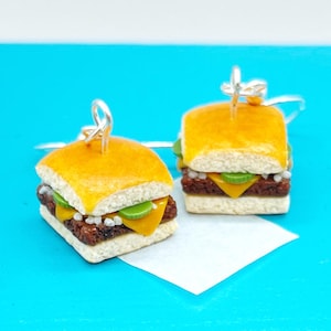May include: A pair of miniature cheeseburger earrings featuring a golden-brown bun, a dark brown patty, a slice of yellow cheese, and green pickle slices. Each burger charm is attached to a silver-toned metal earring hook for hanging.