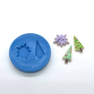 May include: A blue silicone mold with two shapes: a star and a Christmas tree. There are three decorated cookies: a purple star with glitter, and two green Christmas trees with white icing and red sprinkles.