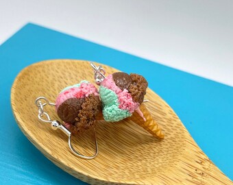Puede incluir: Un par de pendientes con conos de helado en miniatura con bolas de helado rosa, verde y marrón. Los pendientes se muestran en una cuchara de madera sobre un fondo azul.