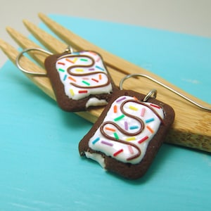Puede incluir: Un par de pendientes con forma de pastelitos cubiertos de chocolate con glaseado blanco y chispas de colores. Los pendientes cuelgan de ganchos plateados.