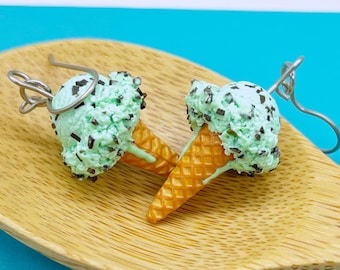 Puede incluir: Un par de pendientes colgantes con forma de conos de helado de menta con virutas de chocolate. Cada pendiente presenta una bola verde claro texturizada con virutas marrón oscuro y una base de cono de barquillo beige, con un gancho de metal plateado.