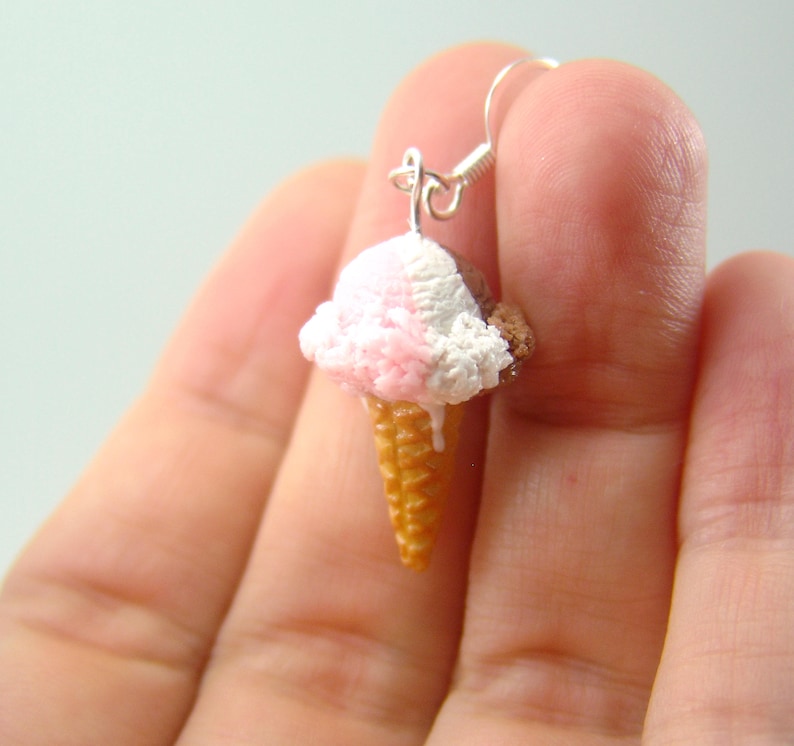 May include: A pair of silver earrings shaped like ice cream cones with pink, white, and brown ice cream. The ice cream is melting down the side of the cone.