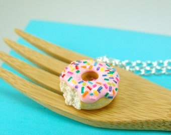 Könnte beinhalten: Miniatur-Donut-Anhänger-Halskette mit einem fehlenden Biss. Der Donut hat rosa Glasur und bunte Streusel. Er liegt auf einer Holzgabel vor blauem Hintergrund, mit einer silbernen Kette sichtbar.