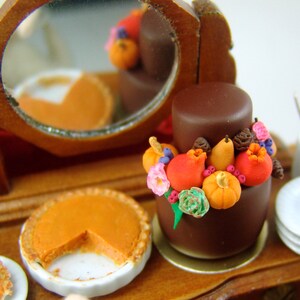 Puede incluir: Un pastel de chocolate en miniatura decorado con acentos de fondant de tem&aacute;tica oto&ntilde;al, incluyendo calabazas, granadas y bayas. El pastel se exhibe en una mesa de madera con un pastelito y un espejo.
