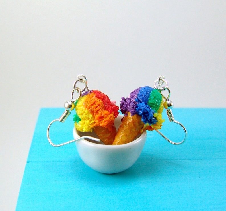 May include: Rainbow ice cream cone earrings with silver hooks. The ice cream is made of polymer clay and is a vibrant mix of red, orange, yellow, green, blue, and purple.