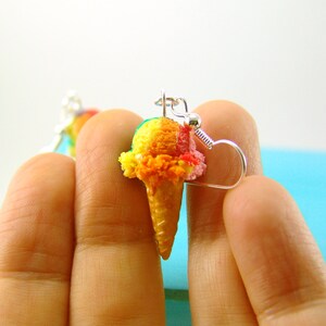 May include: A pair of earrings featuring a miniature ice cream cone with three scoops of colorful ice cream. The ice cream is yellow, orange, and pink. The earrings are made of polymer clay and have silver hooks.