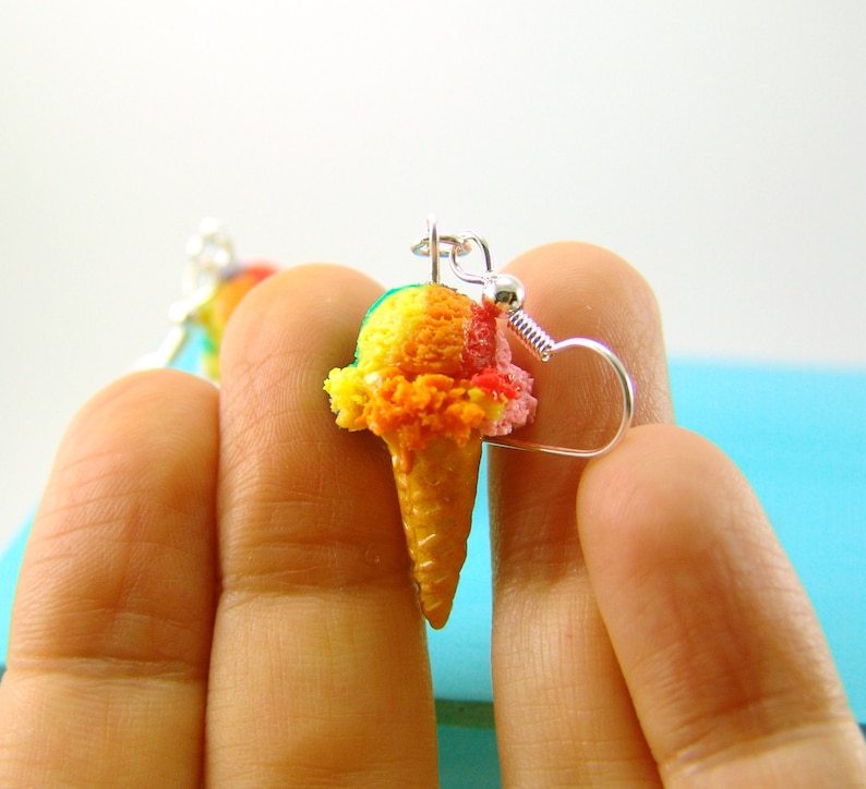 May include: A pair of earrings featuring a miniature ice cream cone with three scoops of colorful ice cream. The ice cream is yellow, orange, and pink. The earrings are made of polymer clay and have silver hooks.