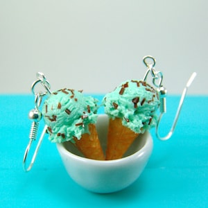 May include: A pair of mint green ice cream cone earrings with chocolate sprinkles. The earrings are displayed in a white bowl on a turquoise background.