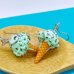 May include: A pair of dangle earrings shaped like mint chocolate chip ice cream cones. Each earring features a textured light green scoop with dark brown chocolate flecks atop a golden-brown waffle cone, attached to a silver-toned metal hook.