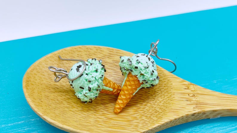 May include: A pair of dangle earrings shaped like mint chocolate chip ice cream cones. Each earring features a textured light green scoop with dark brown chocolate flecks atop a golden-brown waffle cone, attached to a silver-toned metal hook.
