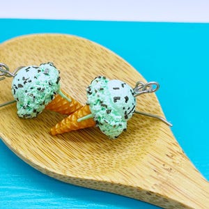May include: A pair of mint chocolate chip ice cream cone earrings. Each earring features a textured light green scoop with dark brown flecks, a golden-brown waffle cone base, and a silver-toned metal jump ring attached to the top for hanging.