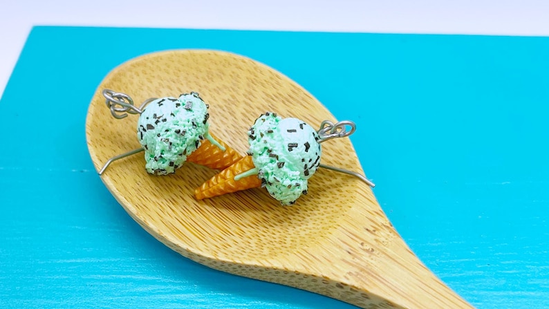 May include: A pair of mint chocolate chip ice cream cone earrings. Each earring features a textured light green scoop with dark brown flecks, a golden-brown waffle cone base, and a silver-toned metal jump ring attached to the top for hanging.